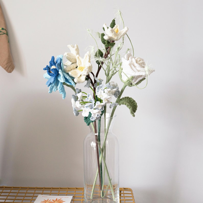 May include: A bouquet of crocheted flowers in a clear glass vase. The flowers are white, blue, and green.