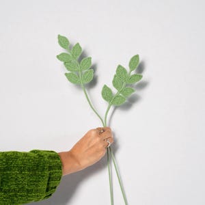 May include: Two stems of crocheted green leaves are held against a white background. The leaves have a detailed texture, and the stems are a lighter shade of green. The leaves are held by a person wearing a green sweater.