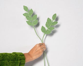 May include: Two stems of crocheted green leaves are held against a white background. The leaves have a detailed texture, and the stems are a lighter shade of green. The leaves are held by a person wearing a green sweater.