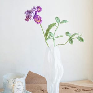 May include: A white, wavy-shaped vase holds a crocheted purple and yellow flower and green leaves. A candle in a glass jar with gold accents sits on a white tray. The candle has the text "Nouvelle Neige".