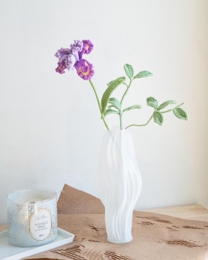 May include: A white, wavy-shaped vase holds a crocheted purple and yellow flower and green leaves. A candle in a glass jar with gold accents sits on a white tray. The candle has the text "Nouvelle Neige".