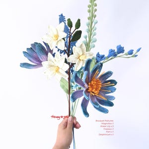 May include: A crocheted bouquet of flowers in shades of blue, white, and green. The bouquet includes a magnolia, two water lilies, a freesia, a fern, and a delphinium. The text "Thenay to you" is written on the stems.