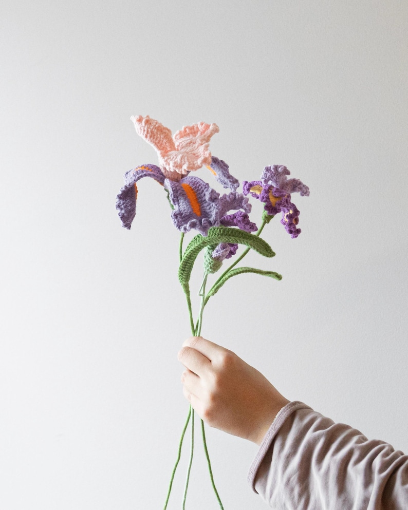 May include: A bouquet of handmade crochet flowers, featuring purple, pink, and orange irises with green stems and leaves. The flowers are held in a person's hand against a plain white background.