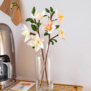 May include: A bouquet of crocheted flowers in shades of white and peach, arranged in a clear glass vase. The flowers are displayed on a gold wire shelf.