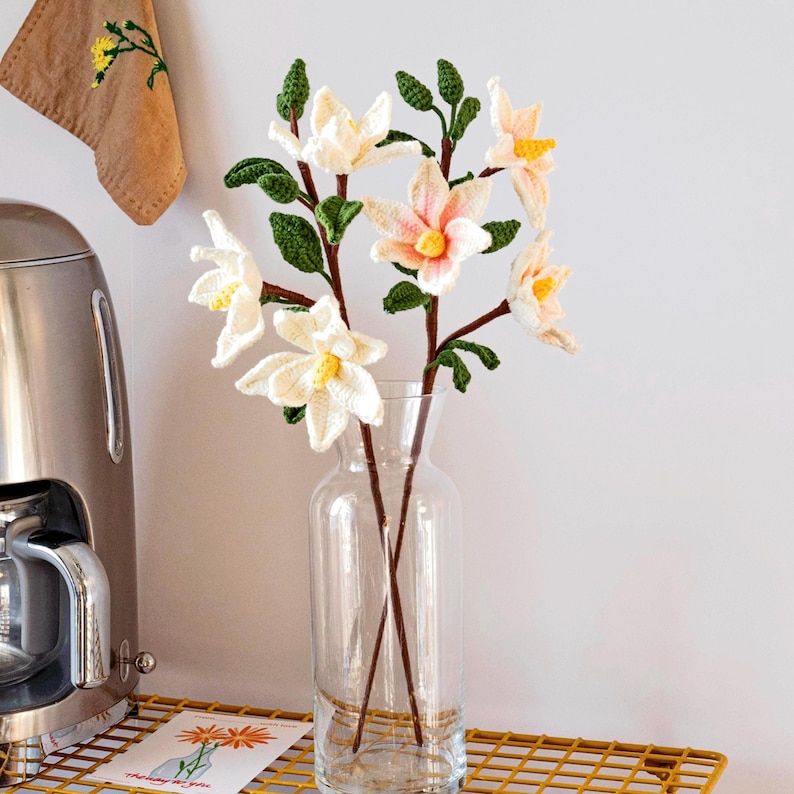 May include: A bouquet of crocheted flowers in shades of white and peach, arranged in a clear glass vase. The flowers are displayed on a gold wire shelf.