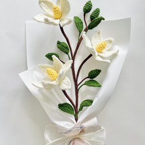 May include: A bouquet of crocheted white magnolia flowers with yellow centres and green leaves, wrapped in white tissue paper and tied with a white ribbon.