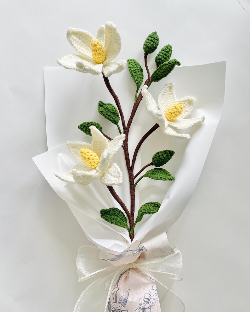 May include: A bouquet of crocheted white magnolia flowers with yellow centres and green leaves, wrapped in white tissue paper and tied with a white ribbon.