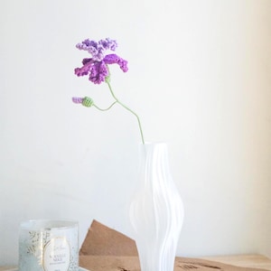 May include: A handmade crochet flower in shades of purple and green, displayed in a white vase. The flower has detailed petals and a delicate stem. A candle in a glass jar and a white tray are also visible.