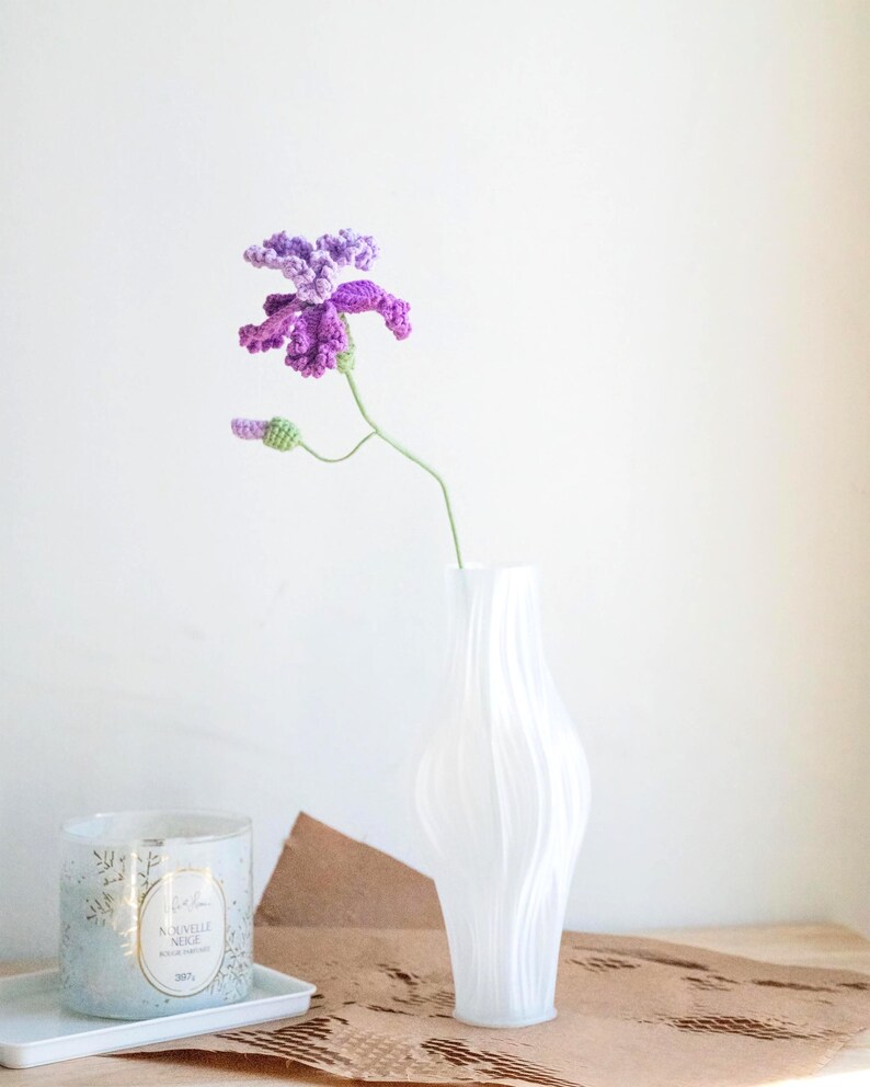 May include: A handmade crochet flower in shades of purple and green, displayed in a white vase. The flower has detailed petals and a delicate stem. A candle in a glass jar and a white tray are also visible.