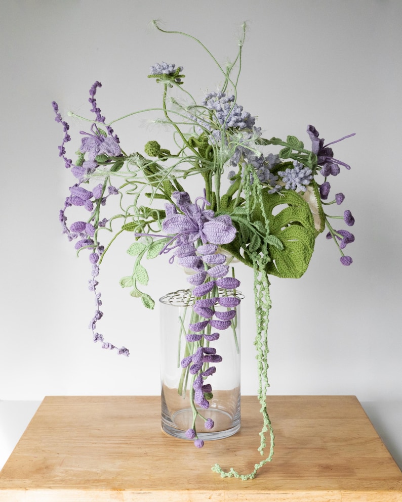 May include: A crocheted bouquet of purple and green flowers in a clear glass vase. The flowers are arranged in a loose, natural style, and the vase is simple and elegant.