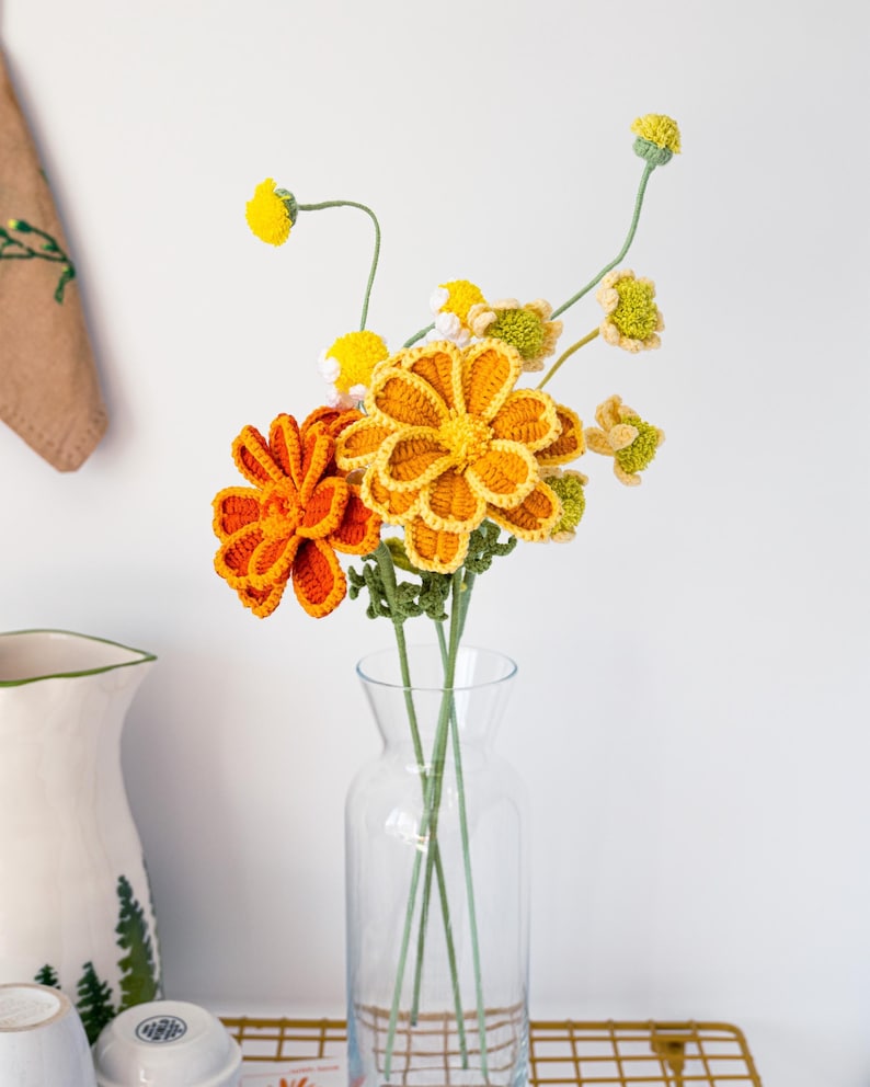 May include: A clear glass vase holds a bouquet of handmade crochet flowers. The flowers are in shades of orange, yellow, and green, with detailed petal designs. The vase sits on a gold-colored wire rack, with a white background.