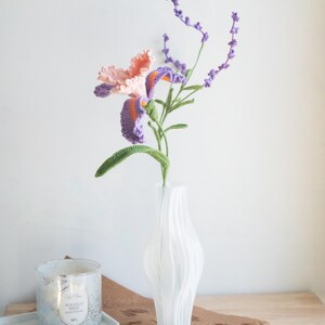 May include: A handmade crochet iris flower with peach and purple petals, orange accents, and green stem, paired with a sprig of lavender. The flower is displayed in a white, textured vase. A candle and tray are in the background.