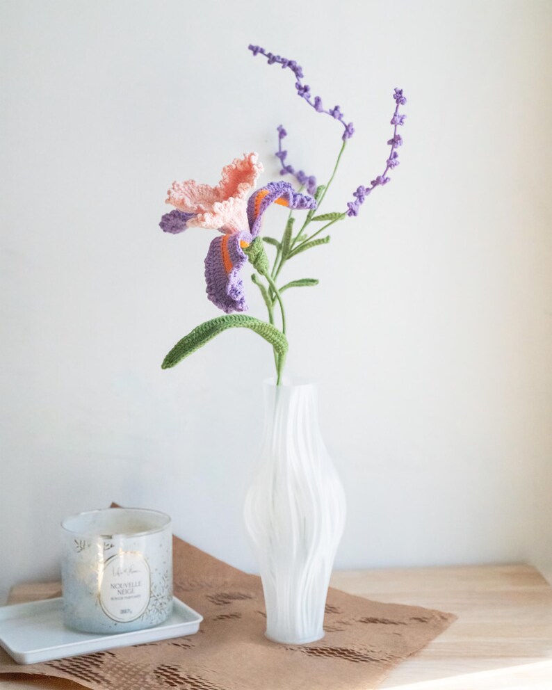 May include: A handmade crochet iris flower with peach and purple petals, orange accents, and green stem, paired with a sprig of lavender. The flower is displayed in a white, textured vase. A candle and tray are in the background.