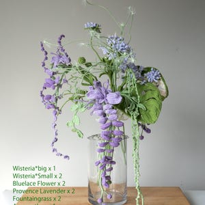 May include: A crocheted flower arrangement in a clear glass vase. The arrangement includes purple wisteria, blue lace flowers, lavender, and green fountaingrass. The flowers are arranged on a wooden surface.