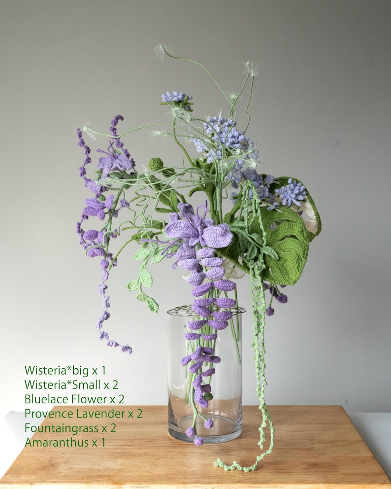 May include: A crocheted flower arrangement in a clear glass vase. The arrangement includes purple wisteria, blue lace flowers, lavender, and green fountaingrass. The flowers are arranged on a wooden surface.