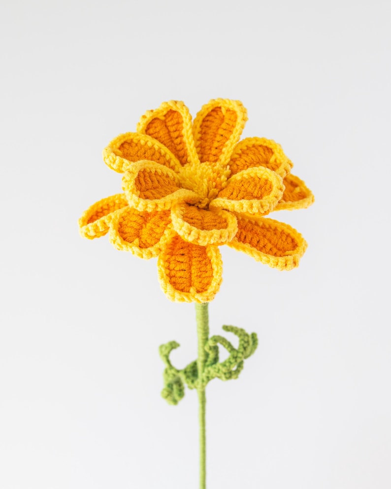 May include: A handcrafted crochet flower with bright yellow petals and a green stem. The flower's petals are intricately detailed, and the stem features small green leaves. The flower is a cheerful, handmade decorative item.