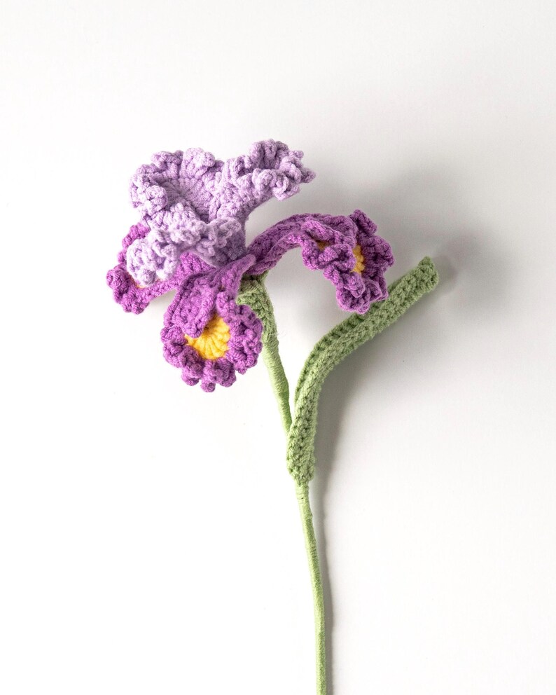 May include: A handcrafted crochet flower with purple petals and a yellow center. The flower has a green stem and leaves. The crochet work is detailed, and the flower is set against a white background.