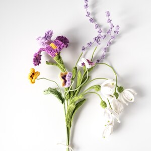 May include: A handmade crochet bouquet featuring a variety of flowers in vibrant colors. The bouquet includes purple irises, yellow and purple pansies, white bell-shaped flowers, and lavender sprigs, all with green stems. The flowers are tied together with a white string.