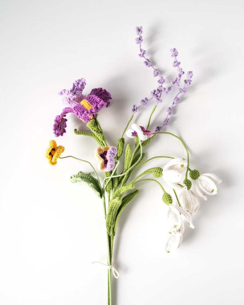 May include: A handmade crochet bouquet featuring a variety of flowers in vibrant colors. The bouquet includes purple irises, yellow and purple pansies, white bell-shaped flowers, and lavender sprigs, all with green stems. The flowers are tied together with a white string.