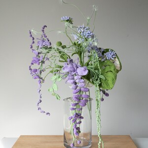 May include: A crocheted bouquet of purple and green flowers in a clear glass vase. The flowers are arranged in a cascading style, with the purple flowers hanging down from the vase.