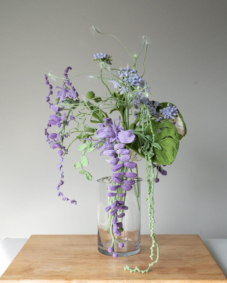 May include: A crocheted bouquet of purple and green flowers in a clear glass vase. The flowers are arranged in a cascading style, with the purple flowers hanging down from the vase.