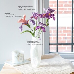 May include: A white vase holds handmade crochet irises in shades of purple, pink, and yellow. The flowers have green stems and are arranged near a window. A candle and a white tray are on a wooden surface. The large iris is 6" x 14" (15cm x 38cm).