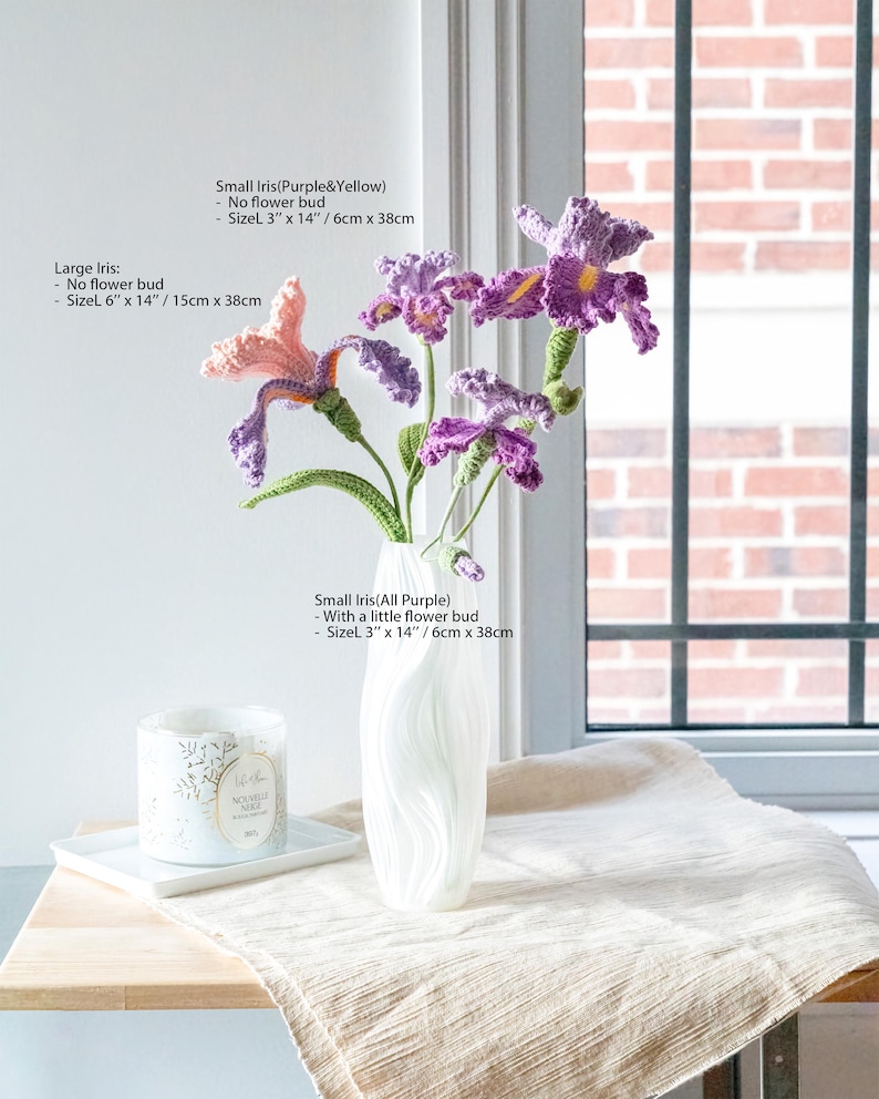 May include: A white vase holds handmade crochet irises in shades of purple, pink, and yellow. The flowers have green stems and are arranged near a window. A candle and a white tray are on a wooden surface. The large iris is 6" x 14" (15cm x 38cm).