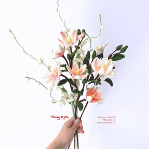 May include: A bouquet of crocheted flowers, including three pink magnolia flowers and two smooth willow branches. The bouquet is held in a hand with the text "the way to you" written below. The bouquet features: Magnolia x 3, Smooth Willow x 2.