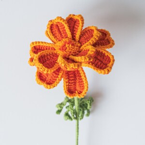 May include: A handmade crochet flower in vibrant orange and yellow hues. The flower features detailed petal work and a textured center, with a green stem and small leaves. The craftwork is set against a plain white background.