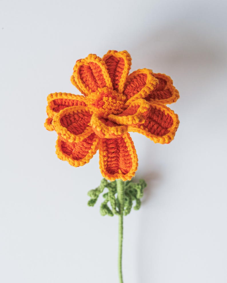 May include: A handmade crochet flower in vibrant orange and yellow hues. The flower features detailed petal work and a textured center, with a green stem and small leaves. The craftwork is set against a plain white background.