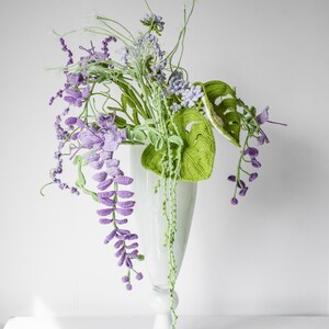 May include: A crocheted bouquet of purple and green flowers in a white vase. The flowers are arranged in a cascading style, with the purple flowers hanging down from the vase.
