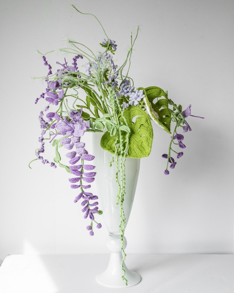 May include: A crocheted bouquet of purple and green flowers in a white vase. The flowers are arranged in a cascading style, with the purple flowers hanging down from the vase.