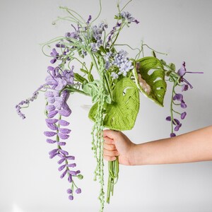 May include: A crocheted bouquet of purple and green flowers. The bouquet includes wisteria, monstera leaves, and other floral elements.