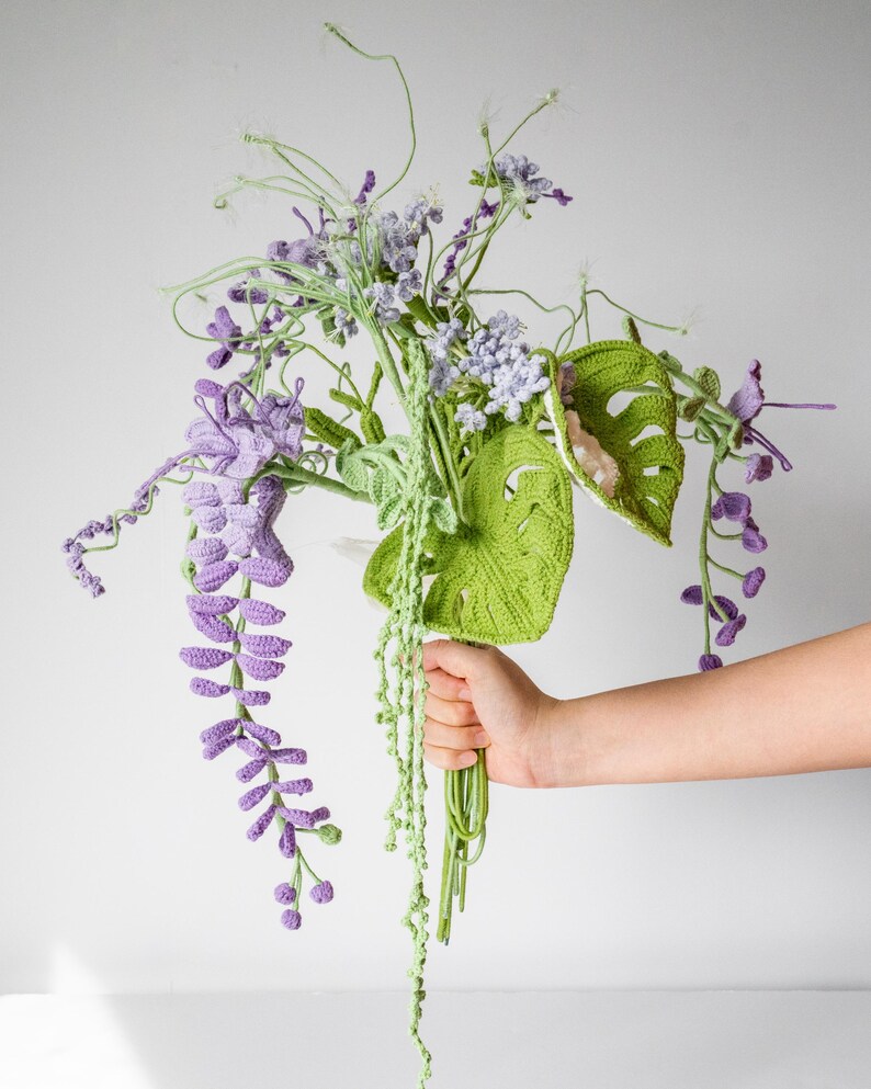 May include: A crocheted bouquet of purple and green flowers. The bouquet includes wisteria, monstera leaves, and other floral elements.