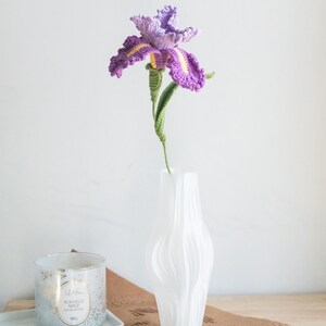 May include: A handmade crochet iris flower in a white vase. The flower has purple petals with yellow accents, a green stem, and is displayed in a textured white vase. A candle in a glass jar sits on a tray in the background.