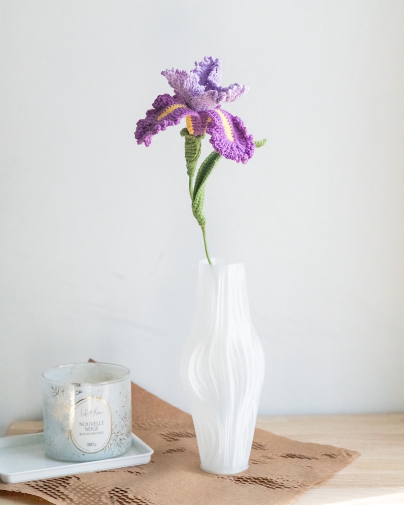 May include: A handmade crochet iris flower in a white vase. The flower has purple petals with yellow accents, a green stem, and is displayed in a textured white vase. A candle in a glass jar sits on a tray in the background.