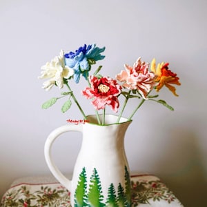 May include: A bouquet of crocheted flowers in a white jug with a green tree design. The flowers are in shades of blue, white, pink, red, and orange.