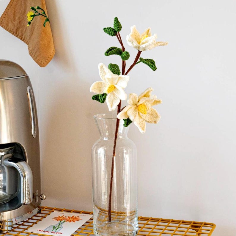 May include: A crocheted white magnolia flower bouquet with green leaves in a clear glass vase on a yellow metal shelf.