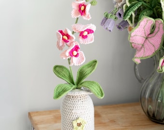 May include: A crocheted orchid plant with pink and fuchsia flowers, green leaves, and a beige vase with a yellow flower accent. The handmade floral arrangement is displayed on a wooden surface.