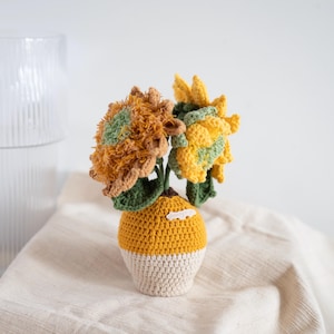 May include: A handmade crochet sunflower arrangement in a yellow and cream vase. The sunflowers have yellow, green, and brown petals. The vase has a small label on the front. The arrangement is on a cream-coloured cloth.