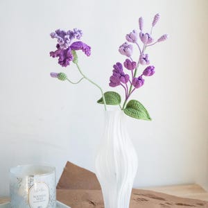 May include: A white, textured vase holds a bouquet of handmade crochet flowers. The flowers are various shades of purple with green leaves and stems. A candle in a glass jar sits to the left of the vase on a white tray.
