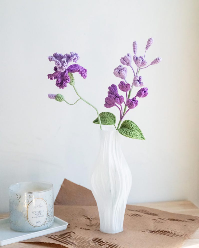 May include: A white, textured vase holds a bouquet of handmade crochet flowers. The flowers are various shades of purple with green leaves and stems. A candle in a glass jar sits to the left of the vase on a white tray.