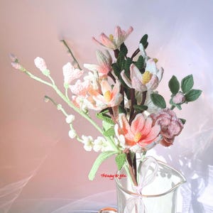 An intricate crochet flower bouquet featuring pastel-toned magnolias with pink and white petals, accompanied by lush green leaves, artistically arranged in a clear glass vase