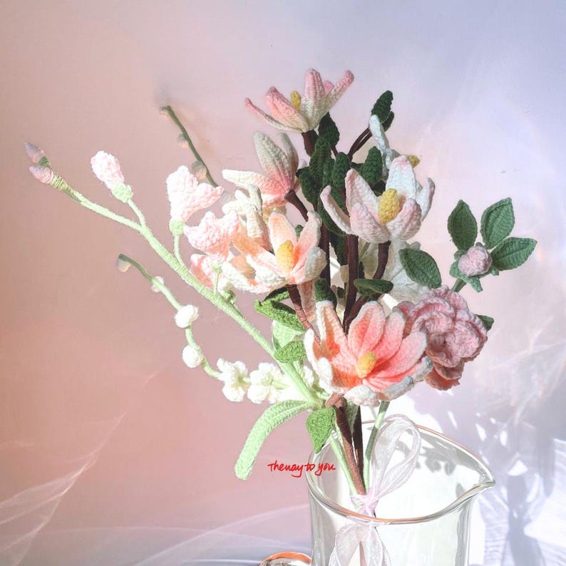 An intricate crochet flower bouquet featuring pastel-toned magnolias with pink and white petals, accompanied by lush green leaves, artistically arranged in a clear glass vase