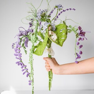 May include: A crocheted bouquet of flowers in shades of green and purple. The bouquet includes a variety of flowers, including wisteria, monstera leaves, and other delicate blooms.