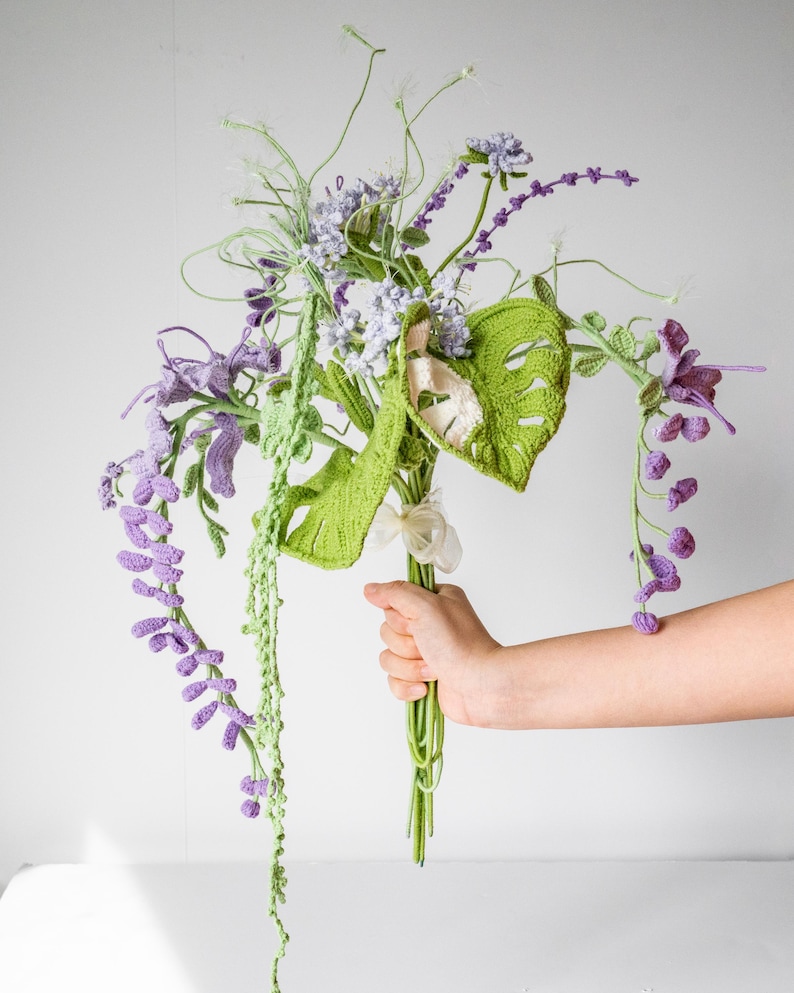May include: A crocheted bouquet of flowers in shades of green and purple. The bouquet includes a variety of flowers, including wisteria, monstera leaves, and other delicate blooms.