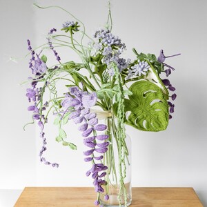 May include: A crocheted bouquet of flowers in a clear glass vase. The bouquet includes purple wisteria, green leaves, and white flowers. The vase is sitting on a wooden table.