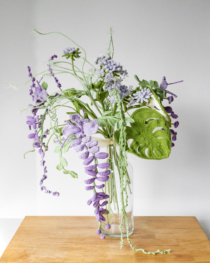 May include: A crocheted bouquet of flowers in a clear glass vase. The bouquet includes purple wisteria, green leaves, and white flowers. The vase is sitting on a wooden table.