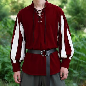 May include: A long-sleeved shirt in deep red with white vertical panels on the sleeves. The shirt features a stand-up collar and a front lace-up closure with black cord. A black leather-like belt with a silver-tone metal ring buckle is worn.