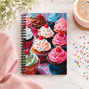 May include: A spiral-bound notebook featuring a colorful illustration of multiple cupcakes with various frosting colors including pink, blue, white, and chocolate, topped with red cherries and multi-colored sprinkles on a dark background.